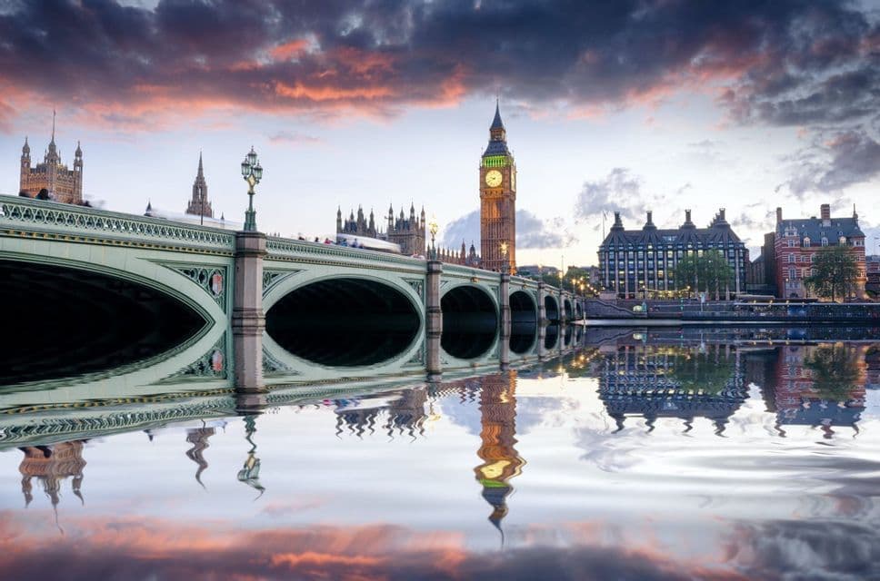 Un ponte ad arco attraversa un fiume calmo con una grande torre dell'orologio e gli edifici della città riflessi nell'acqua al tramonto.
