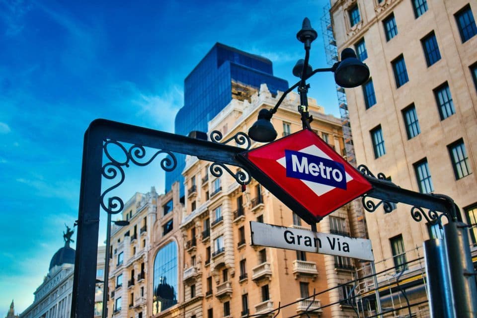Un'insegna d'ingresso di una stazione della metropolitana d'epoca con la scritta Gran Via sullo sfondo di classici edifici cittadini e un cielo blu luminoso.