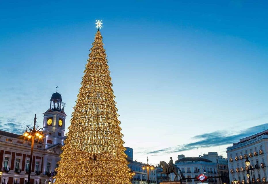 Un alto albero di Natale illuminato si erge in una piazza pubblica della città circondato da edifici storici al crepuscolo