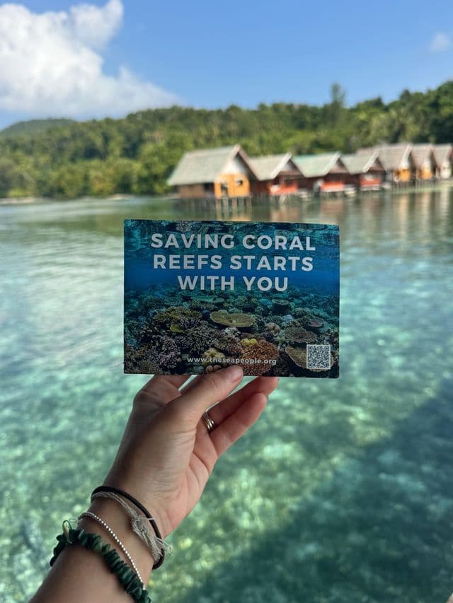 Una mano che tiene una carta per la conservazione della barriera corallina sopra un'acqua turpise limpida con bungalow in legno sullo sfondo
