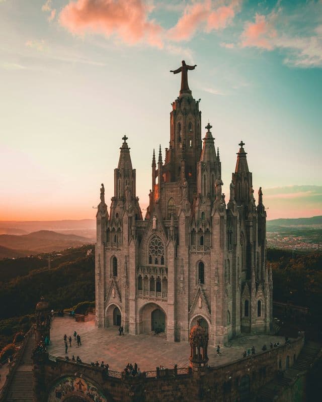 Una iglesia gótica de piedra coronada con una estatua se alza en la colina bajo un cálido cielo al atardecer.
