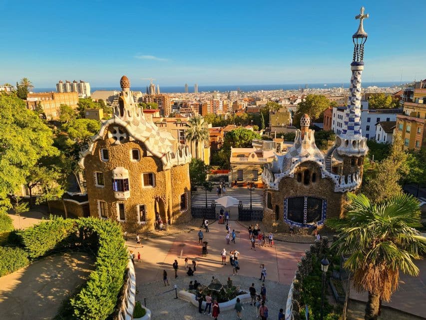 Pabellones de mosaicos ornamentados se encuentran cerca de una plaza de entrada con visitantes, con vistas a una ciudad densa y al mar bajo un cielo despejado.
