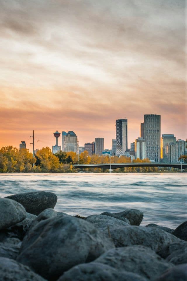 Rocky foreground leading to a flowing river with a modern city skyline and bridge under a sunset sky.