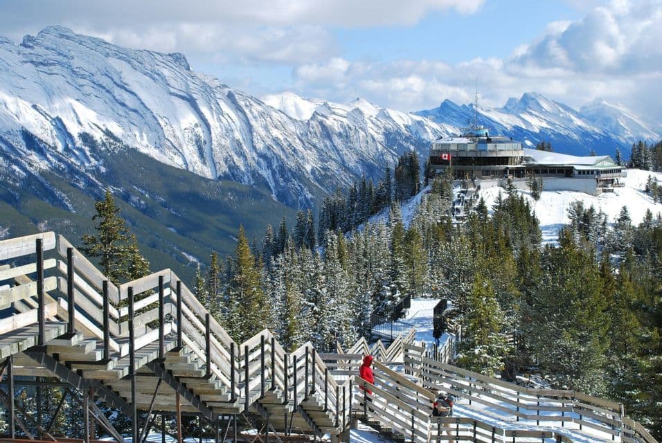 A wooden boardwalk staircase winds along a snowy mountain ridge towards a summit building surrounded by pine trees.