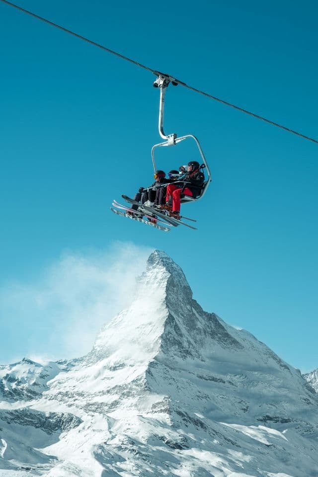 Skiers ride a chairlift high above a snow-covered mountain peak against a clear blue sky.