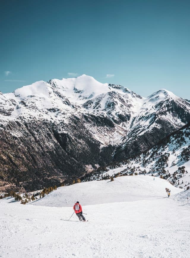 A skier in a red jacket glides down a snowy slope facing massive snow-covered mountain peaks.