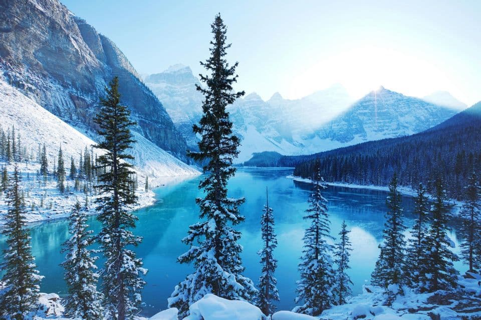 Snow-covered pine trees overlook a bright turquoise lake surrounded by steep, snow-dusted mountains in winter.