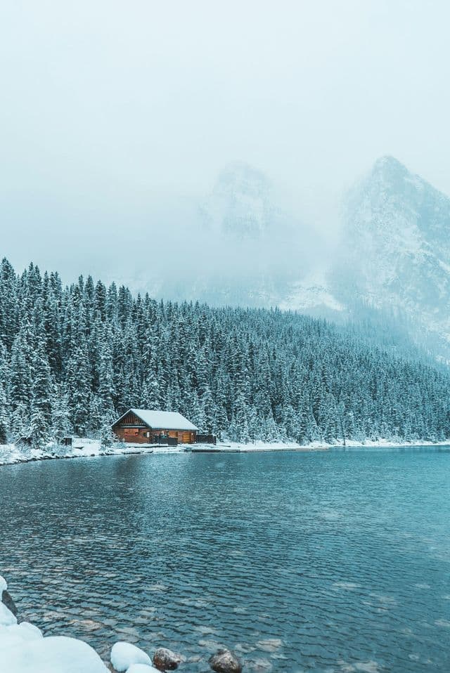 A wooden cabin sits on the shore of a lake surrounded by snow-covered pine forest and misty mountains.