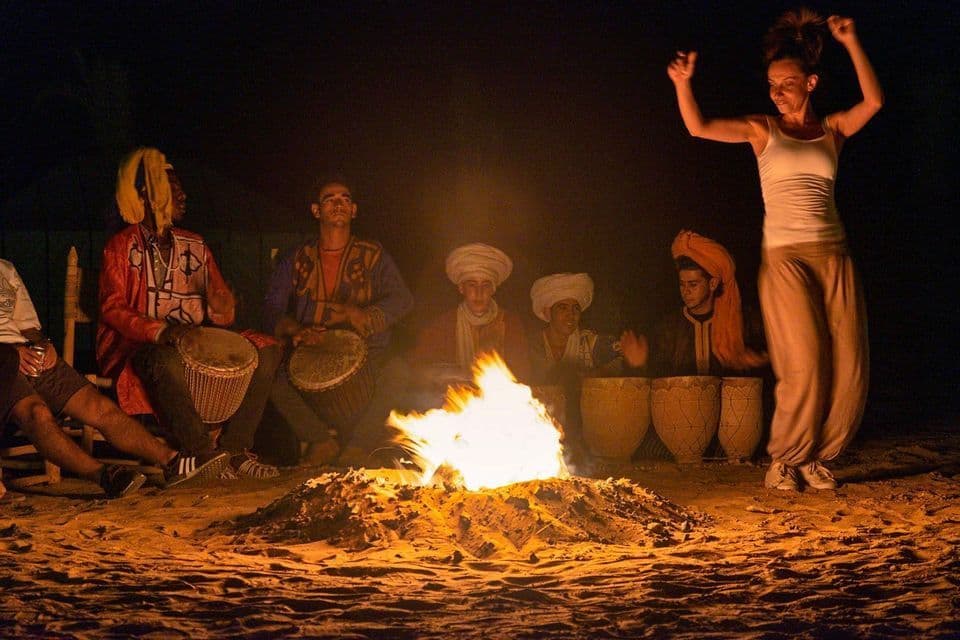 A woman from a WeRoad group trip dances by a bonfire on the sand at night, as men play traditional drums.