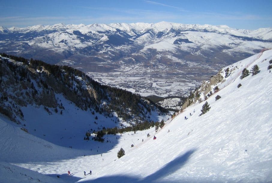Esquiadores descienden por una empinada pendiente nevada con vistas a un valle y picos lejanos bajo un cielo azul.