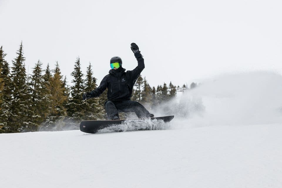 Un snowboarder vestido de negro baja tallando una pendiente nevada, levantando nieve con pinos de fondo