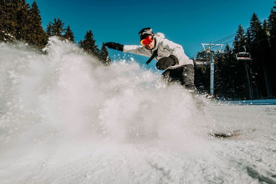 Un snowboarder con gafas levanta nieve polvo mientras baja deslizándose por la pendiente de una montaña cerca de un telesilla.