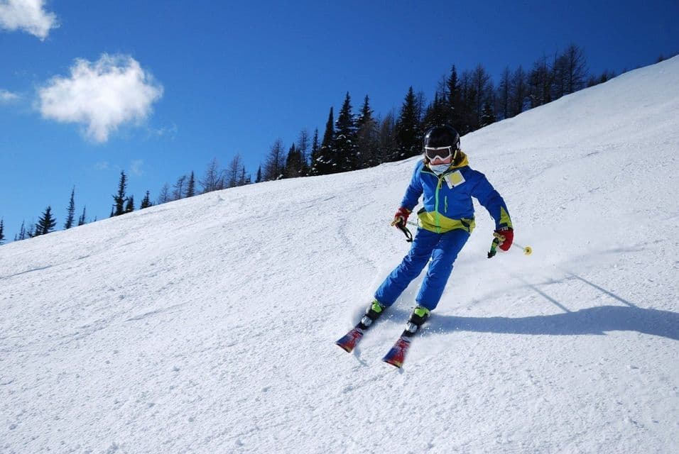 A skier in a blue and yellow suit skiing down a snow-covered slope under a bright blue sky.