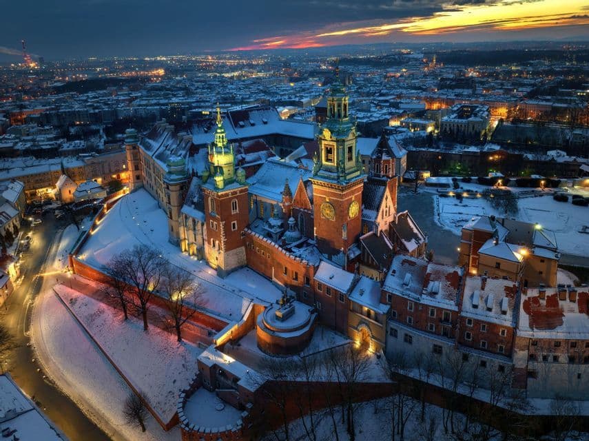 An aerial view of an illuminated historic castle complex covered in snow at dusk overlooking a glowing city skyline.