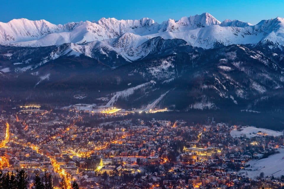 An illuminated town nestled in a valley beneath snow-covered mountain peaks at dusk.