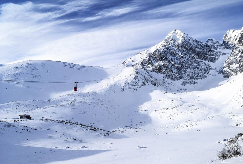 A red cable car travels across snow-covered mountains under a blue sky with wispy clouds.