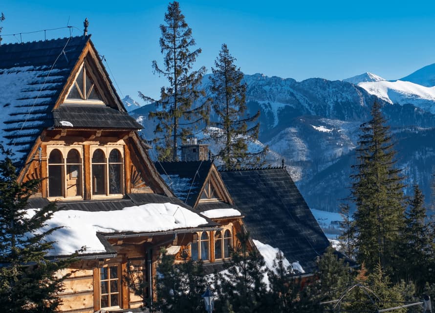 Wooden mountain chalets with snow-covered roofs framed by evergreen pine trees and snowy alpine peaks under a clear blue sky.