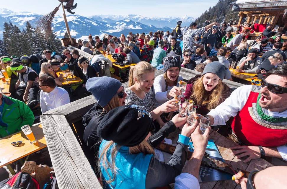 A WeRoad group trip toasting with shot glasses on a crowded sunny terrace overlooking snowy mountains.