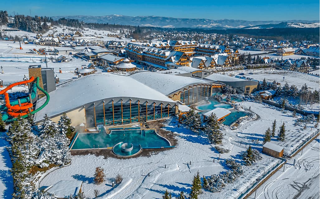 An aerial view of a snow-covered thermal bath resort complex with outdoor steaming pools, water slides, and buildings.