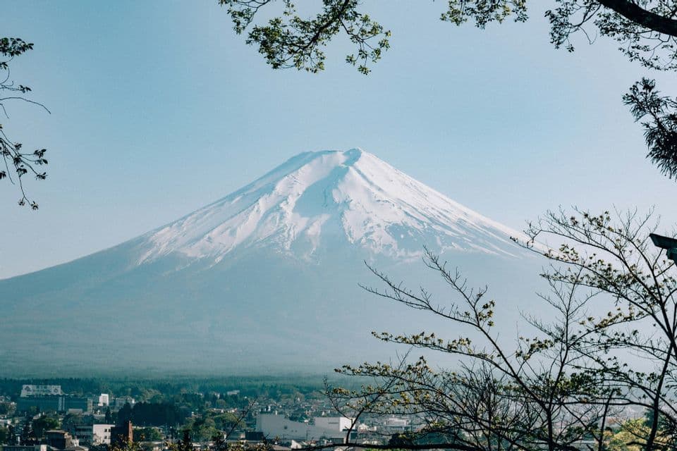 Un grande vulcano innevato si erge sopra una cittadina, incorniciato da rami di alberi verdi sotto un cielo azzurro e limpido.