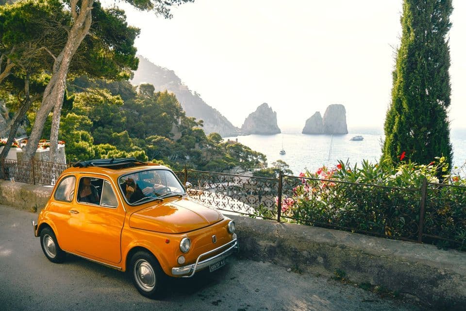 A yellow vintage car parked along a coastal road overlooking sea stacks and boats in the water.