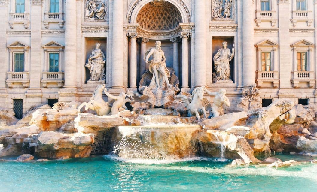 Water cascades into a turquoise pool beneath an ornate classical stone fountain decorated with statues and columns.