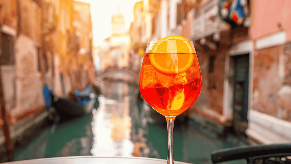 A glass of orange spritz cocktail with an orange slice sitting on a table with a canal and boats in the background.