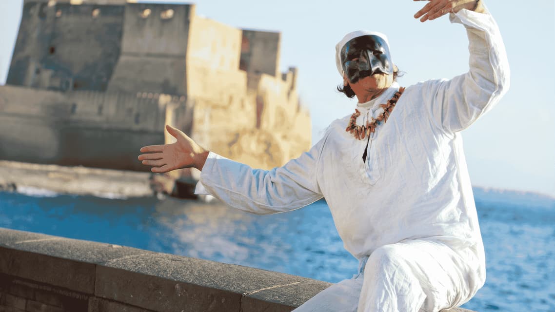 A performer in a traditional Pulcinella costume and black mask gestures dynamically beside a coastal stone wall.