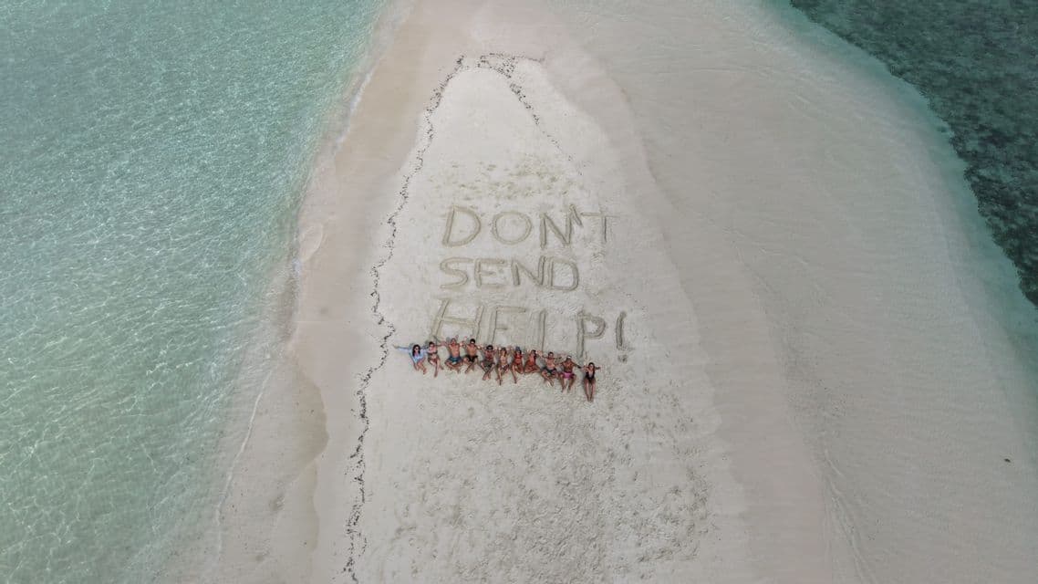 Un viaggio di gruppo WeRoad sdraiati su un banco di sabbia accanto alla scritta 'NON MANDATE AIUTO!' tracciata nella sabbia e circondata da acqua cristallina.