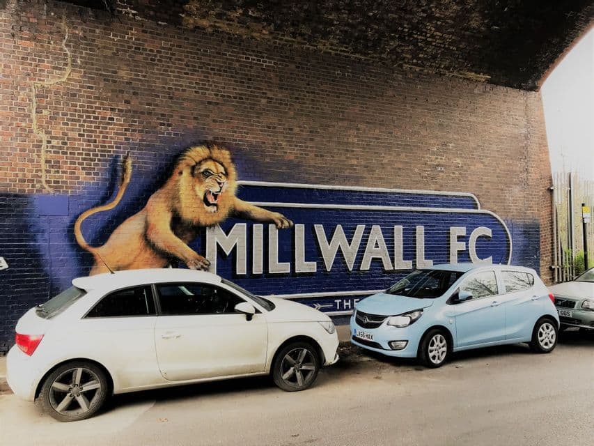 Auto parcheggiate lungo il muro di un sottopassaggio in mattoni dipinto con un murale del Millwall FC raffigurante un leone ruggente.