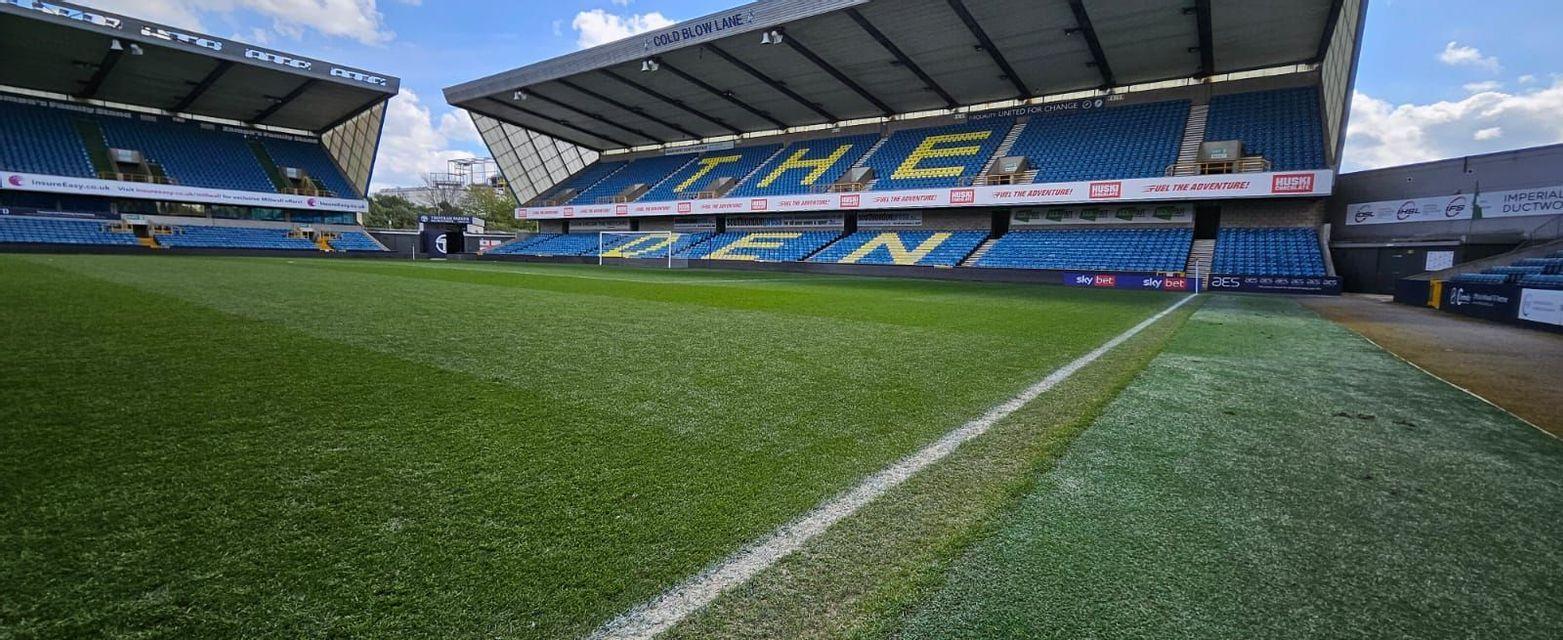 Un campo da calcio vuoto con prato verde, linee di campo bianche e spalti blu sotto un cielo parzialmente nuvoloso
