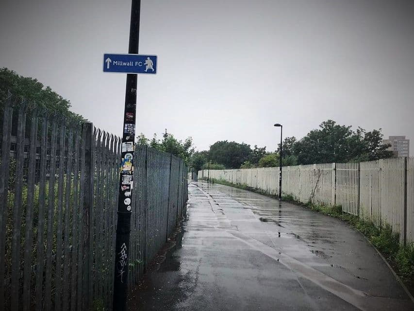 Un sentiero pedonale bagnato affiancato da alte recinzioni metalliche con un cartello di indicazione su un palo in una giornata nuvolosa