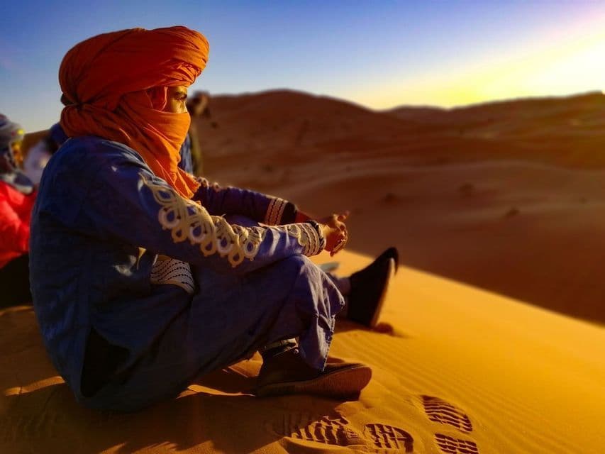 A person on a WeRoad group trip, wearing an orange turban, sits on a sand dune watching the desert sunset.