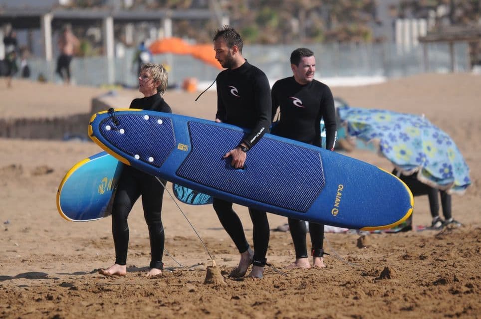 Un viaje en grupo de WeRoad con tres personas en trajes de neopreno llevando tablas de surf y caminando por una playa de arena.