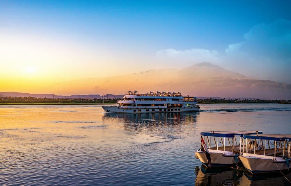 Una grande nave da crociera naviga lungo un ampio fiume al tramonto, con barche più piccole ormeggiate vicino alla riva.