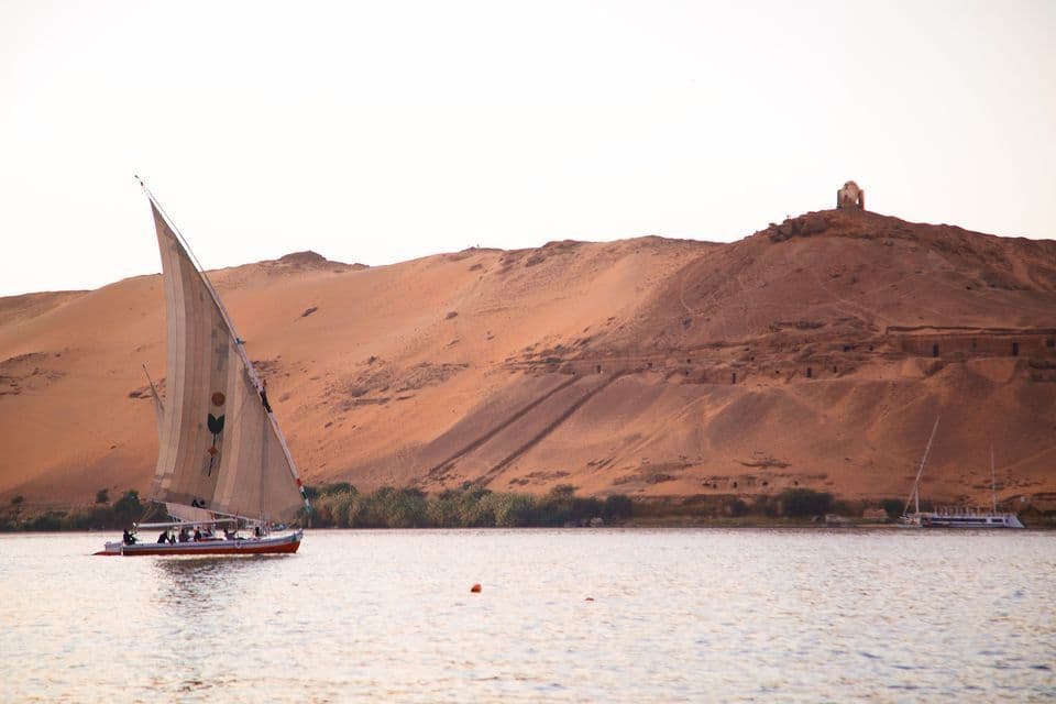 Una barca a vela tradizionale naviga su un ampio fiume sullo sfondo di colline desertiche aride e dune.