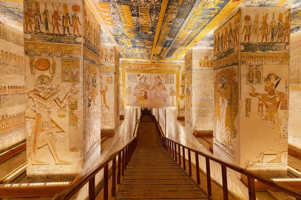 Una passerella di legno scende in un antico corridoio di una tomba circondato da colonne e soffitti decorati con geroglifici.