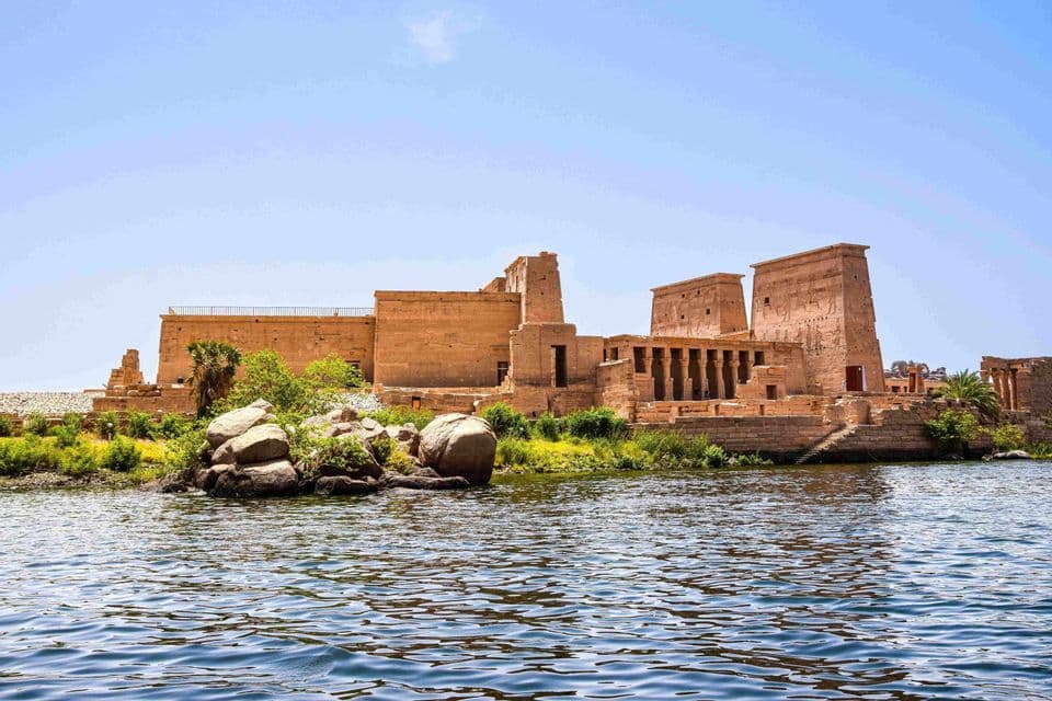 Un antico complesso di templi in pietra arenaria si erge su un'isola rocciosa lungo uno specchio d'acqua sotto un cielo luminoso.