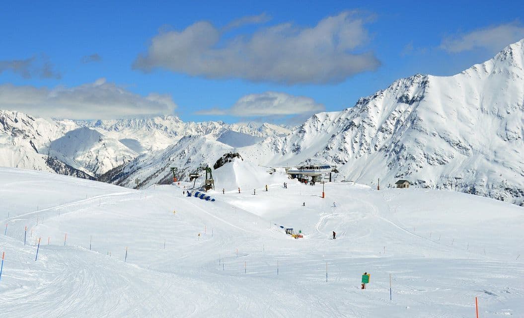 Sciatori e impianti di risalita su piste innevate circondate da alte cime montuose sotto un cielo azzurro e luminoso.