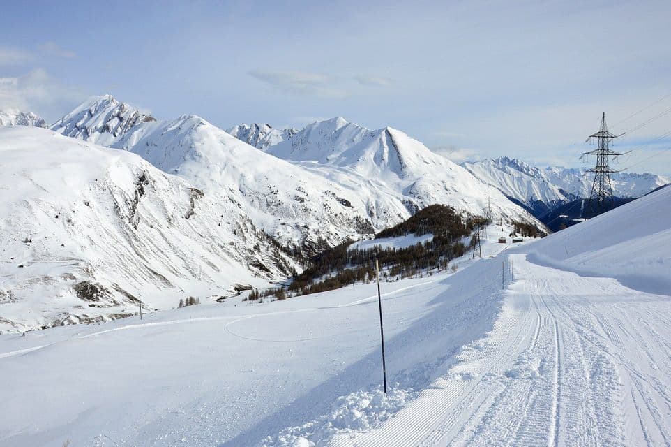 Una pista da sci battuta si snoda lungo creste montuose innevate sotto un cielo limpido con linee elettriche in lontananza