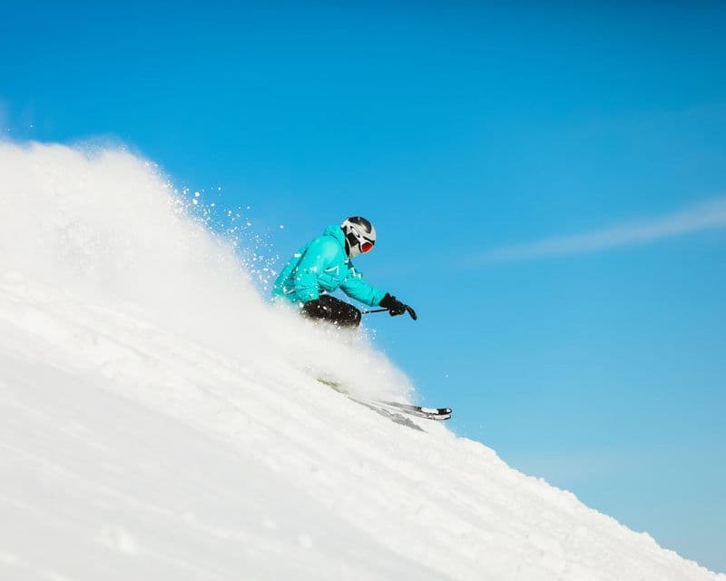Un sciatore con giacca turchese e casco sfreccia su una pista innevata sollevando neve fresca sotto un cielo blu