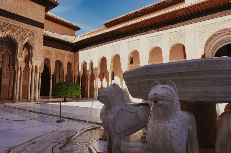 Una fuente de piedra sostenida por estatuas de leones esculpidas que arrojan agua en un patio ornamentado con arcos detallados.