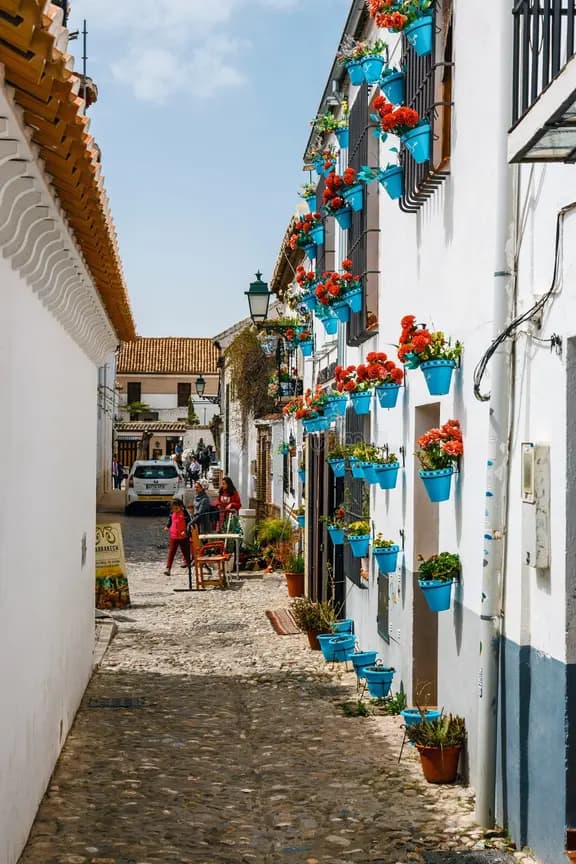 Una estrecha calle empedrada bordeada de edificios encalados decorados con coloridas macetas de flores colgando en las paredes.