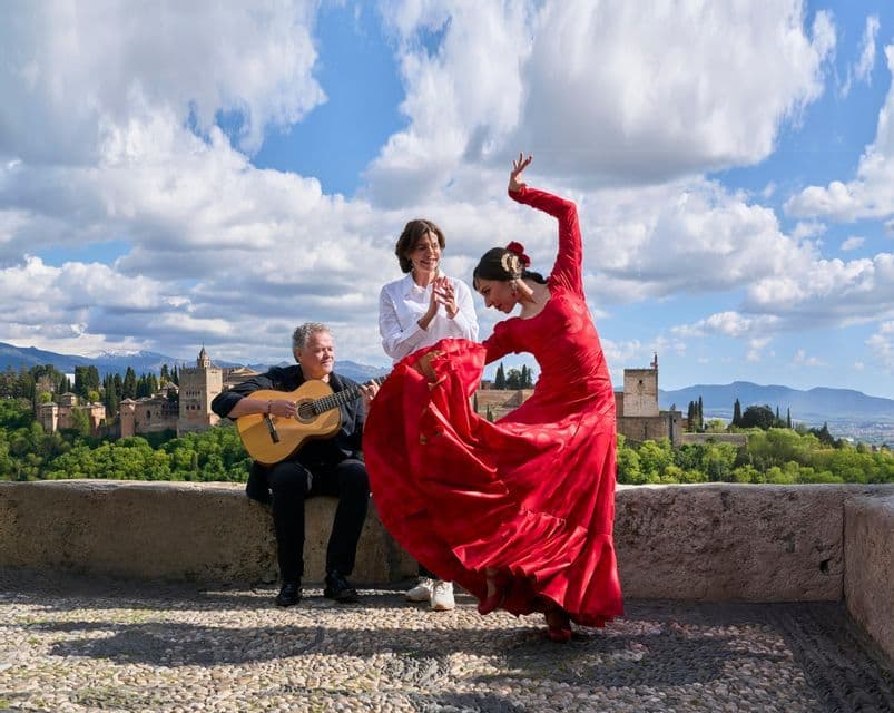 Una bailarina con vestido rojo interpreta flamenco junto a un guitarrista acústico y una mujer tocando las palmas en una terraza de piedra al aire libre.
