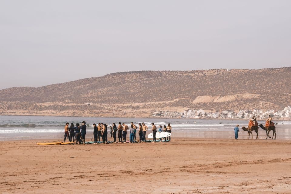 Un viaggio di gruppo WeRoad si riunisce con letavole da surf su un'ampia spiaggia di sabbia vicino all'oceano e ai cammelli