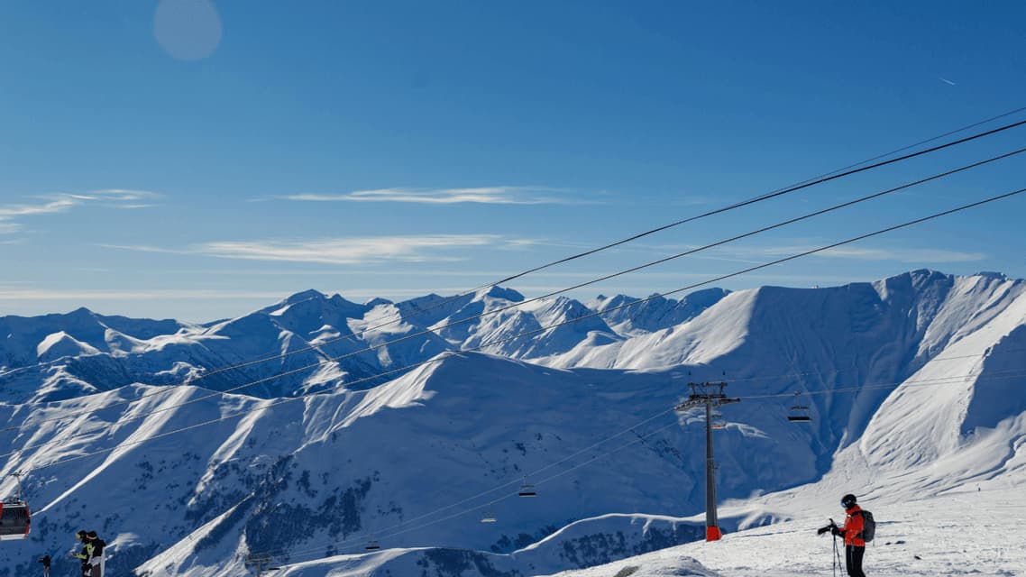 Un sciatore si trova su un pendio innevato vicino agli impianti di risalita che dominano vaste catene montuose innevate sotto un cielo limpido.