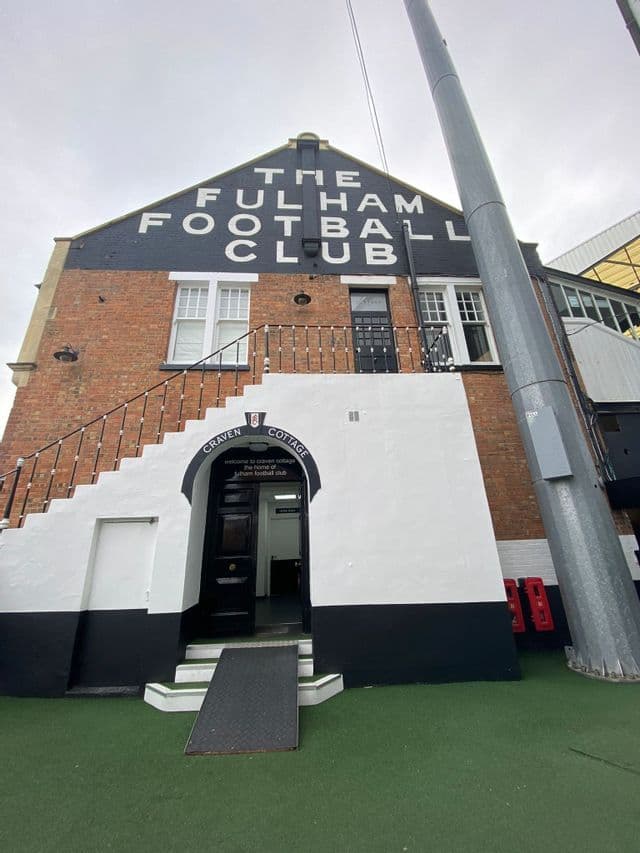 Edificio di uno stadio in mattoni con un ingresso ad arco con la scritta Craven Cottage e una scala esterna accanto al prato verde.