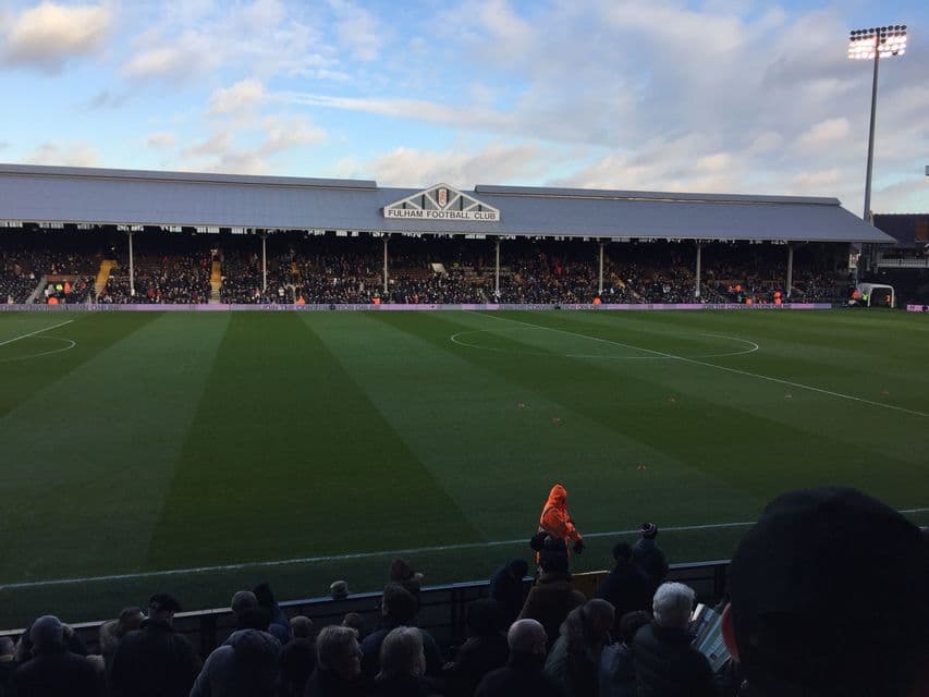 Spettatori seduti in una tribuna coperta dello stadio che si affaccia su un campo da calcio verde sotto un cielo parzialmente nuvoloso