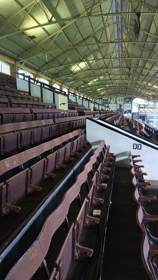 File di sedili da stadio in legno d'epoca sotto un tetto a capriate metalliche ad arco all'interno di una tribuna sportiva vuota.