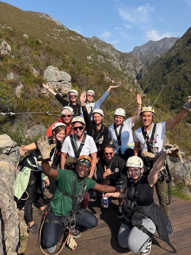 Eine WeRoad-Gruppe posiert gemeinsam mit Helmen und Zipline-Gurtzeugen auf einer Holzplattform inmitten von Felsbergen.